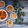 Aerial view of assorted nuts and seeds in white bowls with green leaves on a rustic wooden table.