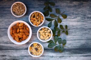 Aerial view of assorted nuts and seeds in white bowls with green leaves on a rustic wooden table.